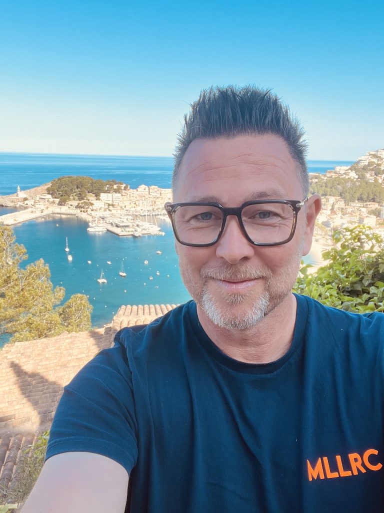 AiSLAND - Gastgeber Marc Ulrich Eine Person (Marc Ulrich) mit Brille und Bart lächelt in die Kamera. Im Hintergrund ist eine malerische Küstenstadt mit Bucht, Booten und strahlend blauem Himmel zu sehen. Im Vordergrund sind Bäume und Dächer zu sehen.