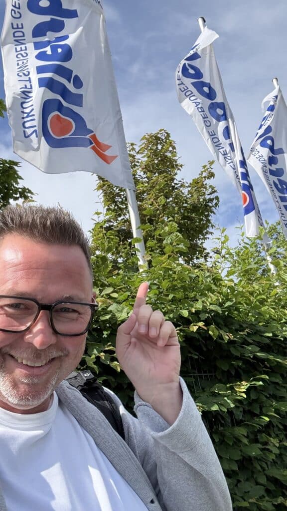 Ein lächelnder Mann mit Brille zeigt im Freien auf drei weiße Fahnen mit blauen und orangefarbenen Logos.