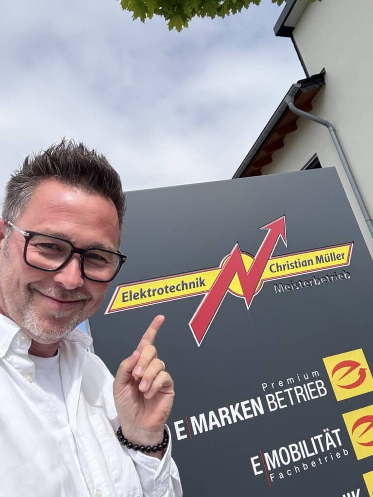 Ein Mann zeigt im Freien auf das Firmenschild von „Elektrotechnik Christian Müller Meisterbetrieb“.