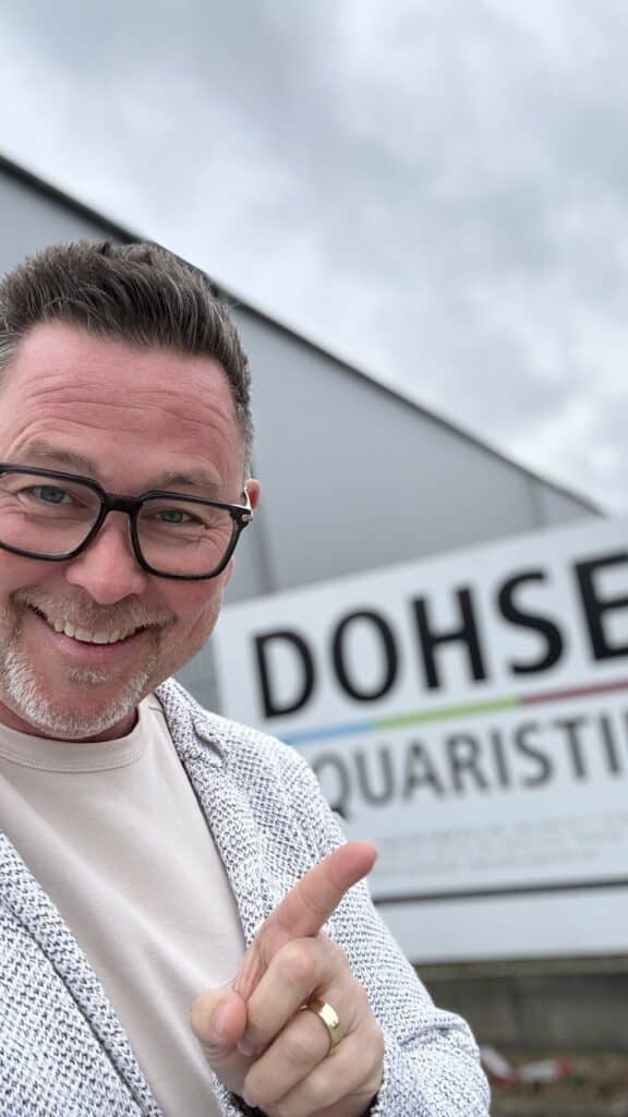 Ein Mann mit Brille zeigt auf ein Schild mit der Aufschrift „DOHSE AQUARISTIK“ und lächelt.