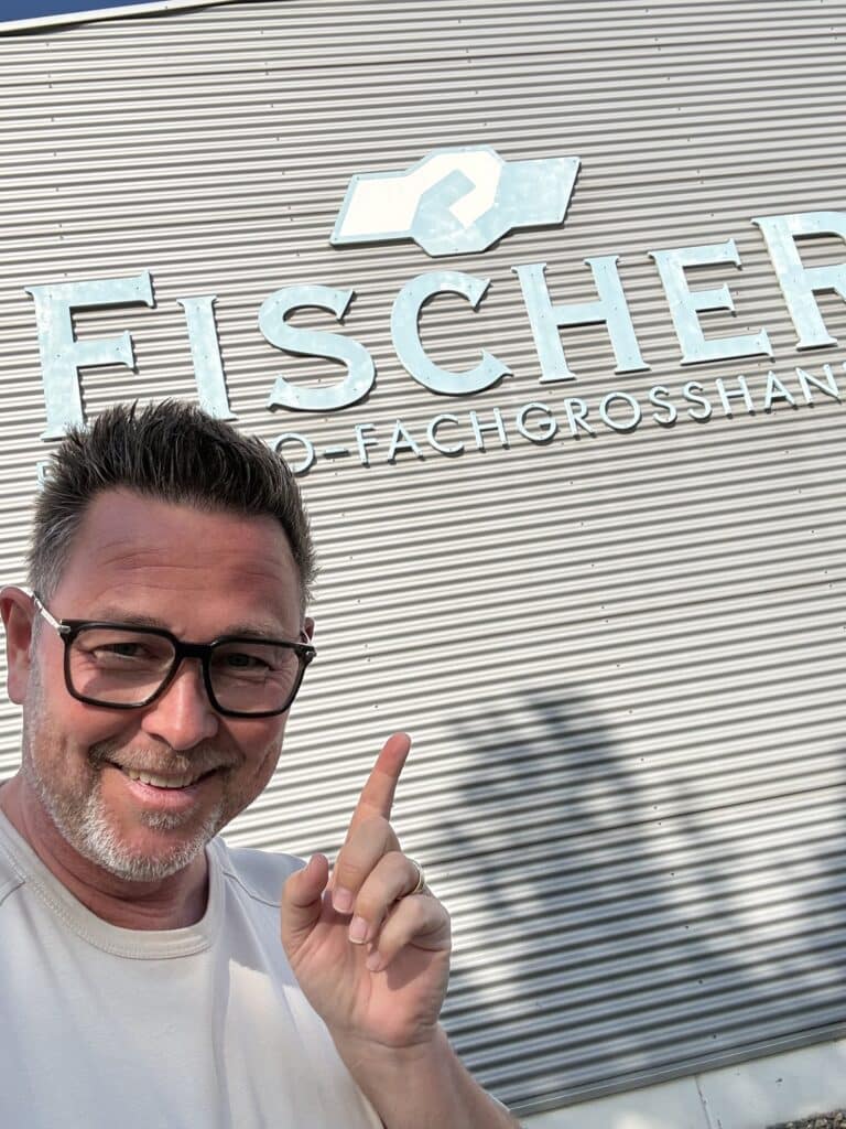 Ein Mann mit Brille zeigt auf ein Gebäudeschild, auf dem in großen Buchstaben „Fischer“ steht.