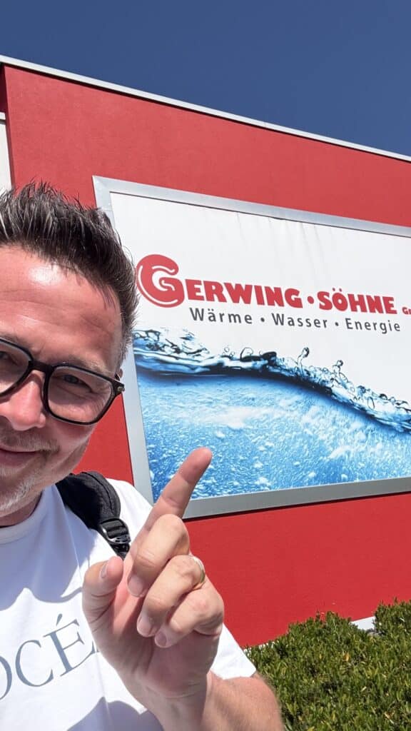 Ein Mann zeigt auf das Firmenschild von Gerwing Söhne an einer roten Wand mit einer Wassergrafik.