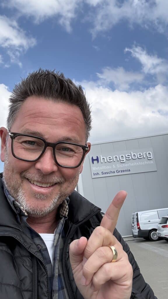 Ein Mann lächelt und zeigt auf ein Gebäude mit der Aufschrift „Hengsberg Metallbau & Sicherheitstechnik“.