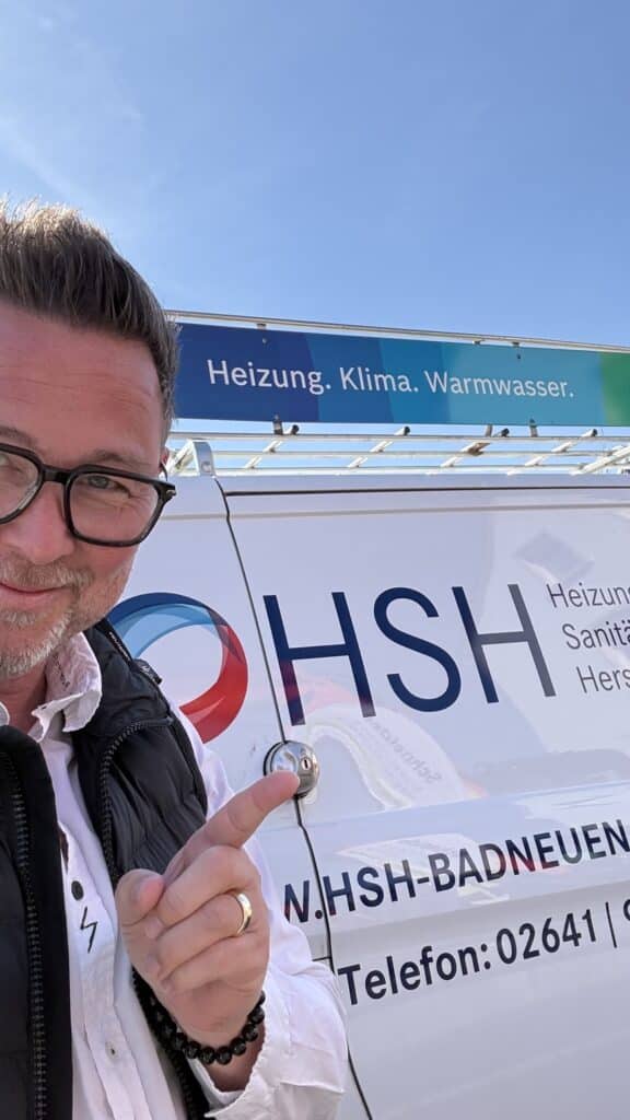 Ein Mann zeigt auf einen Firmenwagen mit dem HSH-Logo und einem Schild mit der Aufschrift „Heizung. Klima. Warmwasser.“.