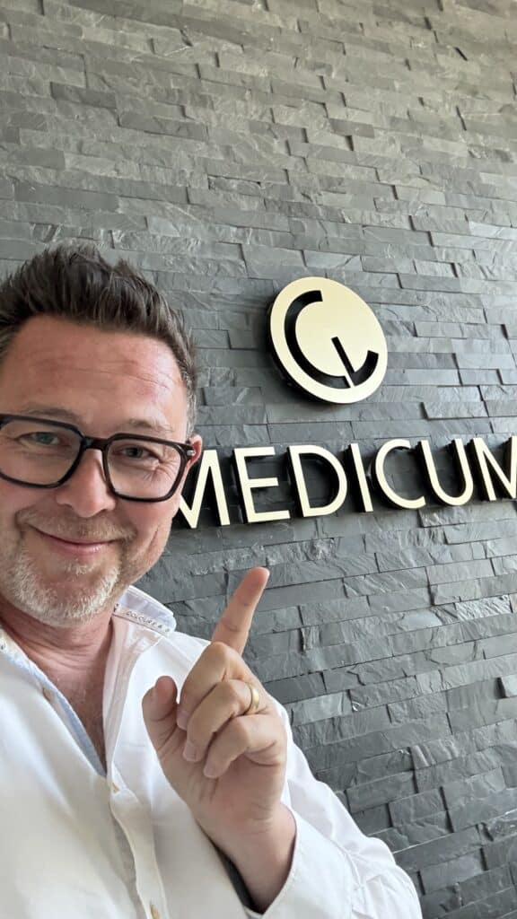 Ein Mann mit Brille zeigt auf ein Wandschild, auf dem vor einem grauen Steinhintergrund „MEDICUM“ steht.