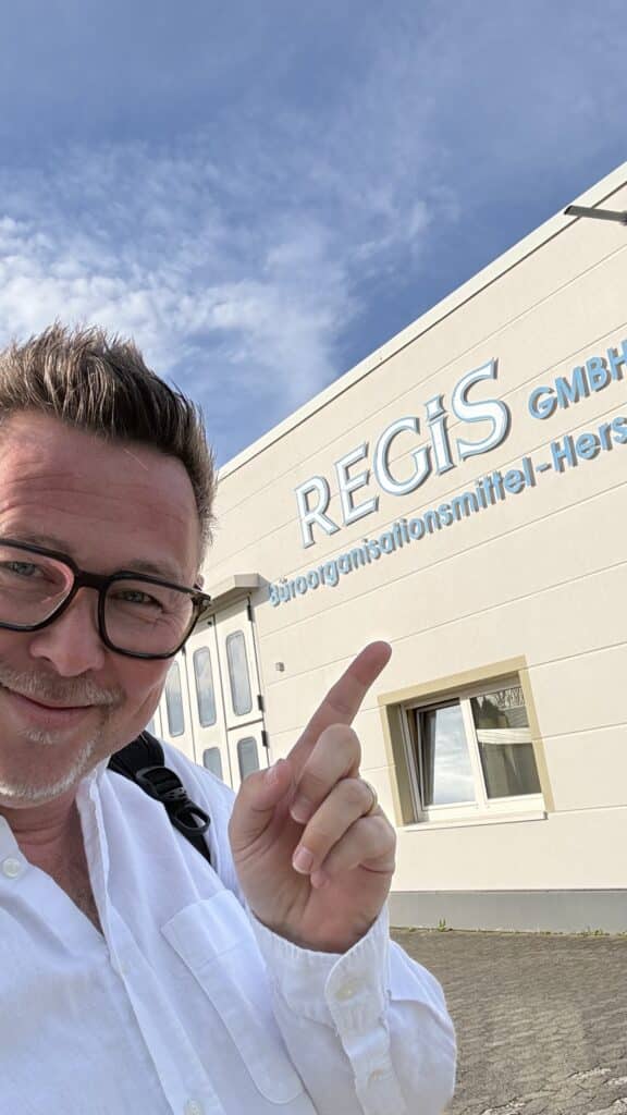 Ein Mann mit Brille zeigt unter blauem Himmel auf ein Gebäude, an dem ein Schild mit der Aufschrift „REGIS GmbH“ hängt.